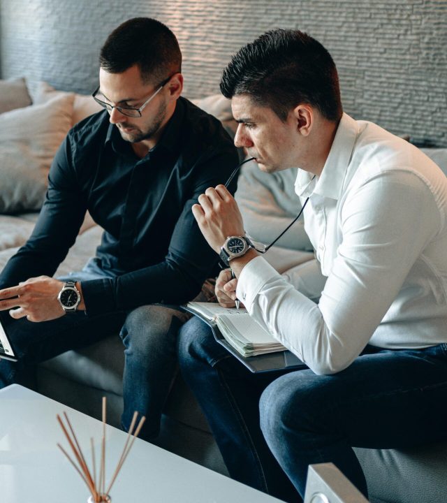 Two men discussing financial data on a tablet while sitting indoors.